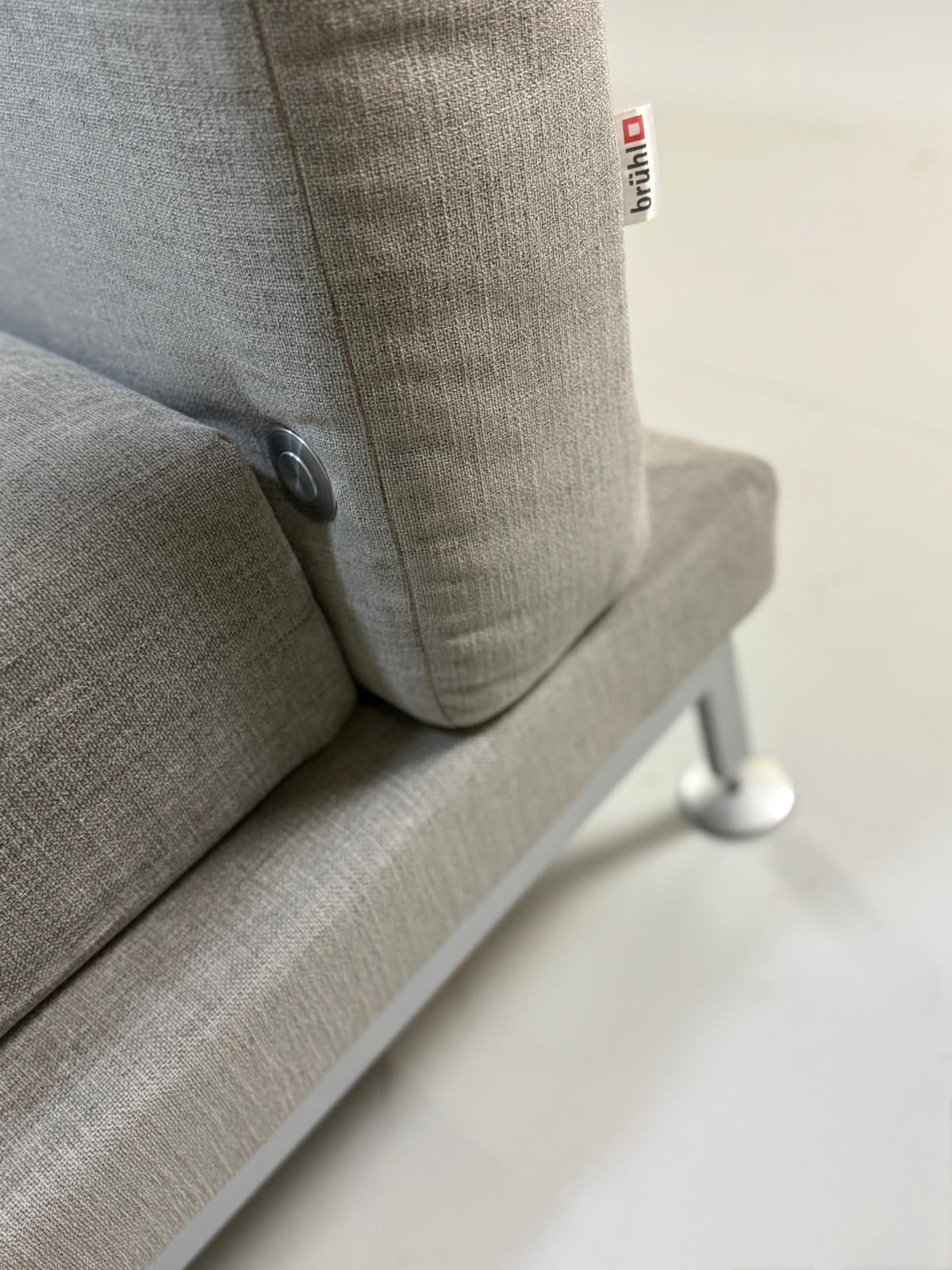 Gebrauchtes graues Stoffsofa Brühl Modell Moule vor einer modernen Wand.