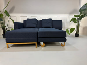 Gebrauchtes blaues Stoffsofa Brühl Modell Fourtwo vor einer modernen Wand.