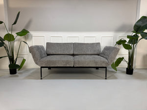 Gebrauchtes graues Stoffsofa Brühl Modell Roro Spring vor einer modernen Wand.