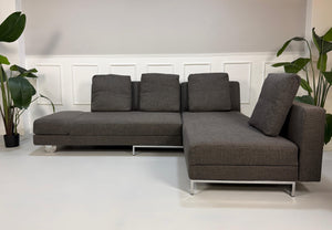 Gebrauchtes graues Brühl Fourtwo Designer Stoff Ecksofa vor einer Wand, stilvoll und modern eingerichtet.