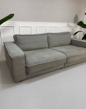 Gebrauchtes graues Bolia Noora Designer Stoff Sofa vor einer Wand, stilvoll und modern eingerichtet.