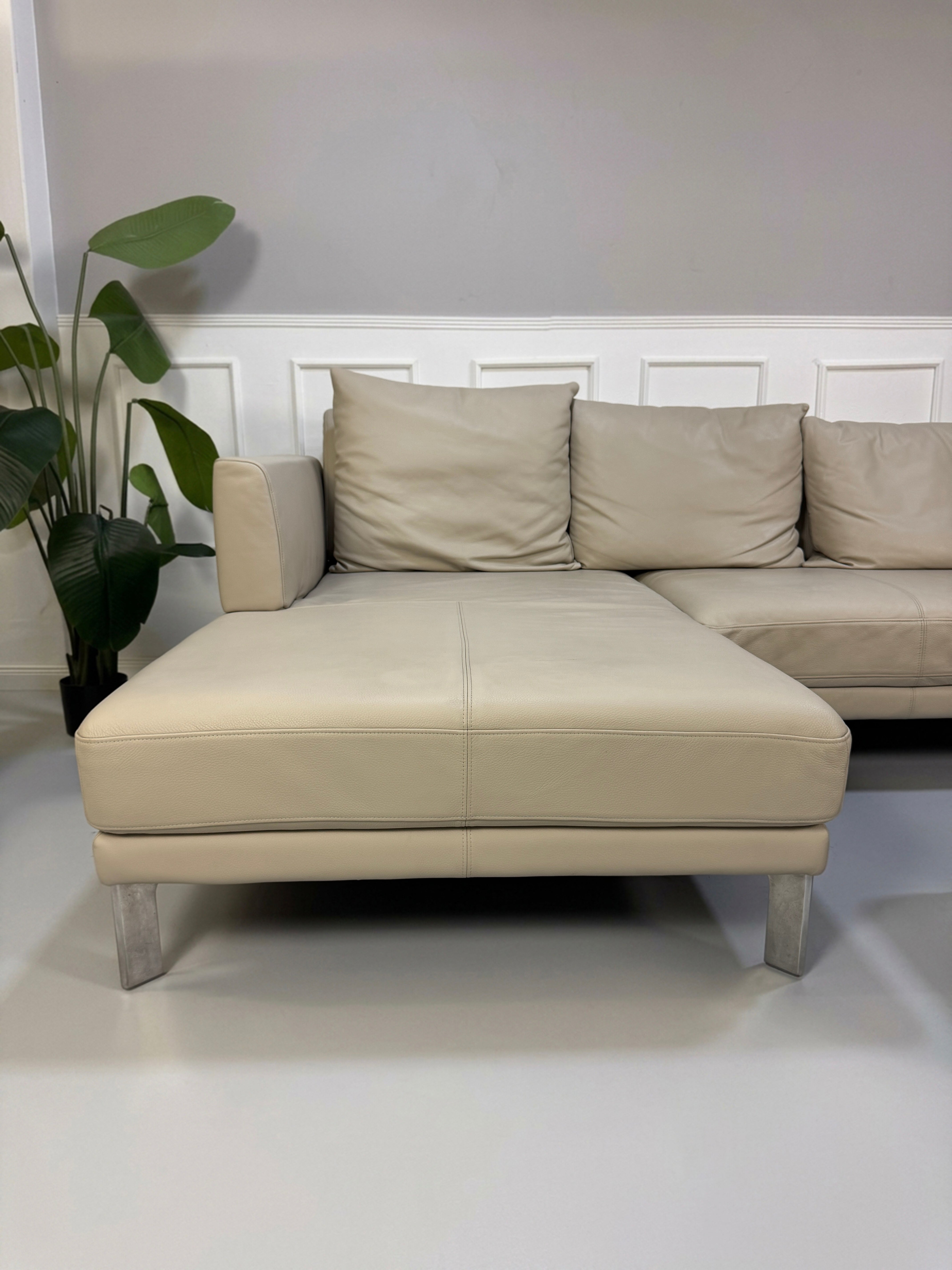 Gebrauchtes beiges Rolf Benz Sento 344 Designer Leder Ecksofa mit Hockerbank vor einer Wand, stilvoll und modern eingerichtet.