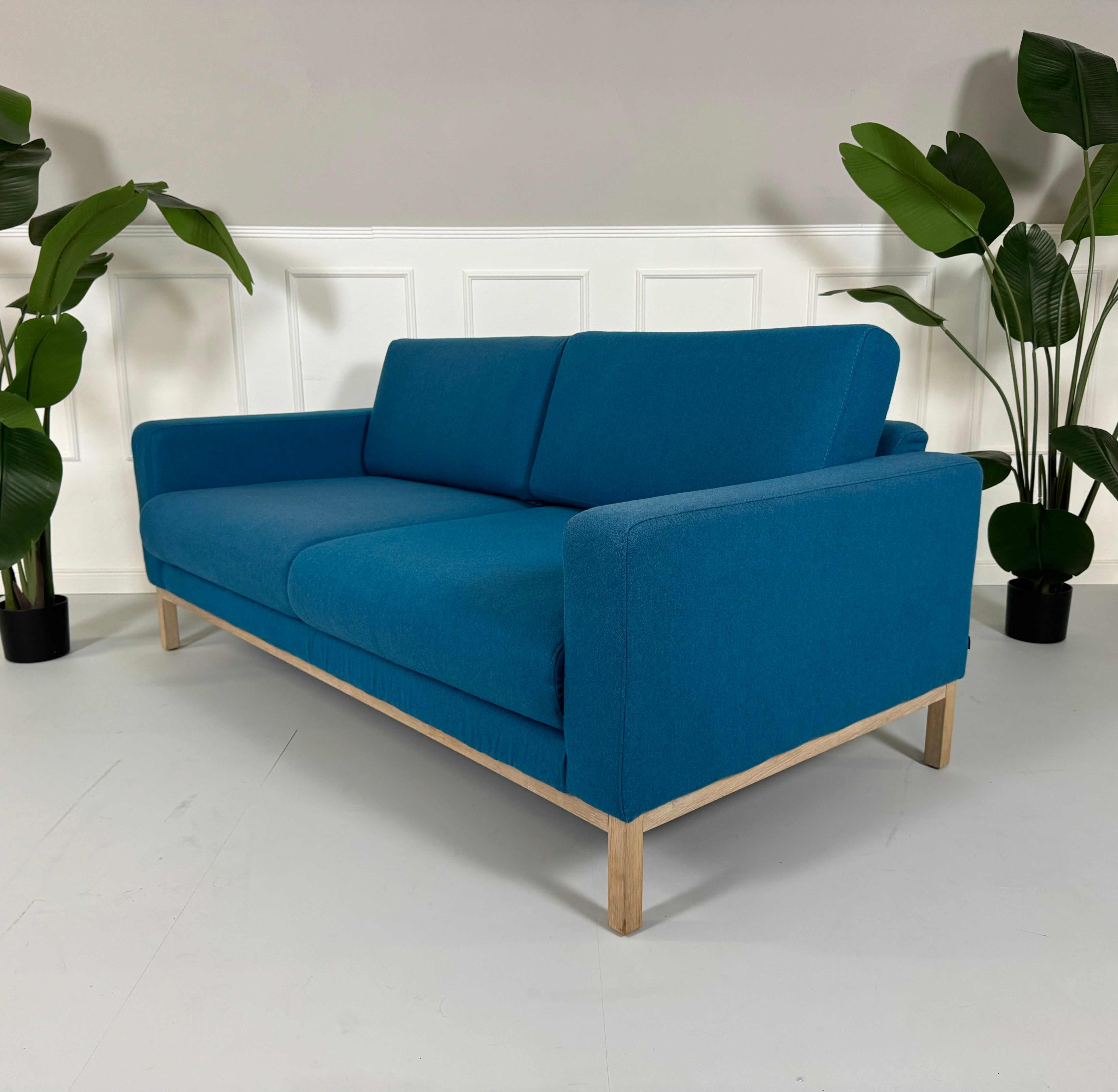 Gebrauchtes blaues petrol Bolia North Designer Stoff Sofa vor einer Wand, stilvoll und modern eingerichtet.