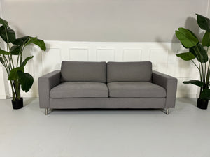Gebrauchtes graues Bolia Scandinavia Stoff Sofa vor einer Wand, stilvoll und modern eingerichtet.