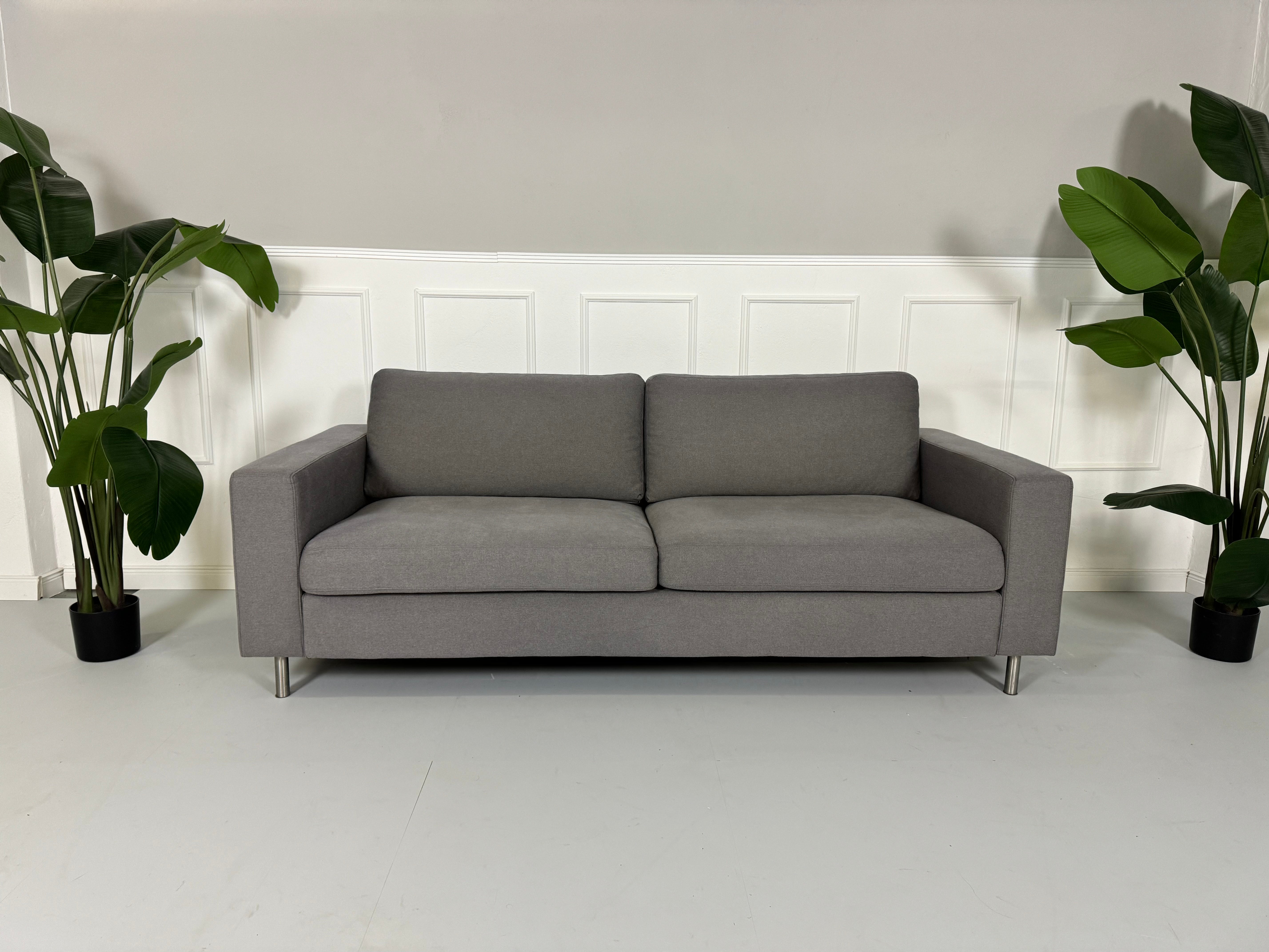 Gebrauchtes graues Bolia Scandinavia Stoff Sofa vor einer Wand, stilvoll und modern eingerichtet.