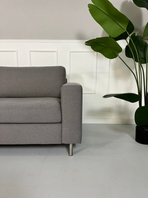 Gebrauchtes graues Bolia Scandinavia Stoff Sofa vor einer Wand, stilvoll und modern eingerichtet.