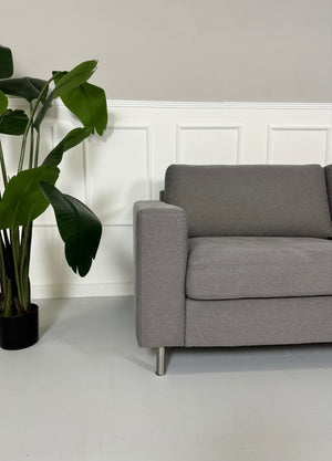 Gebrauchtes graues Bolia Scandinavia Stoff Sofa vor einer Wand, stilvoll und modern eingerichtet.