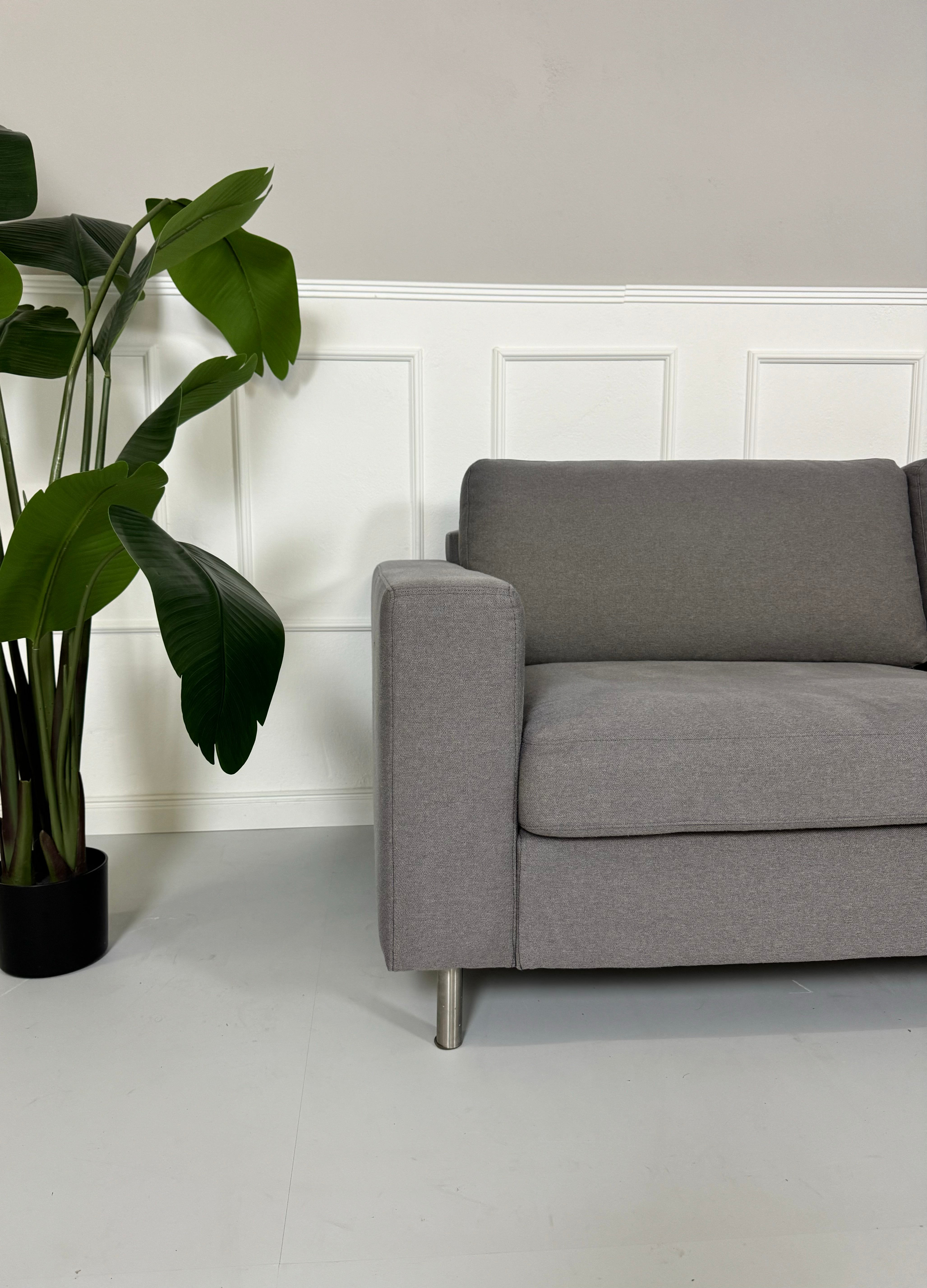 Gebrauchtes graues Bolia Scandinavia Stoff Sofa vor einer Wand, stilvoll und modern eingerichtet.