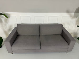 Gebrauchtes graues Bolia Scandinavia Stoff Sofa vor einer Wand, stilvoll und modern eingerichtet.