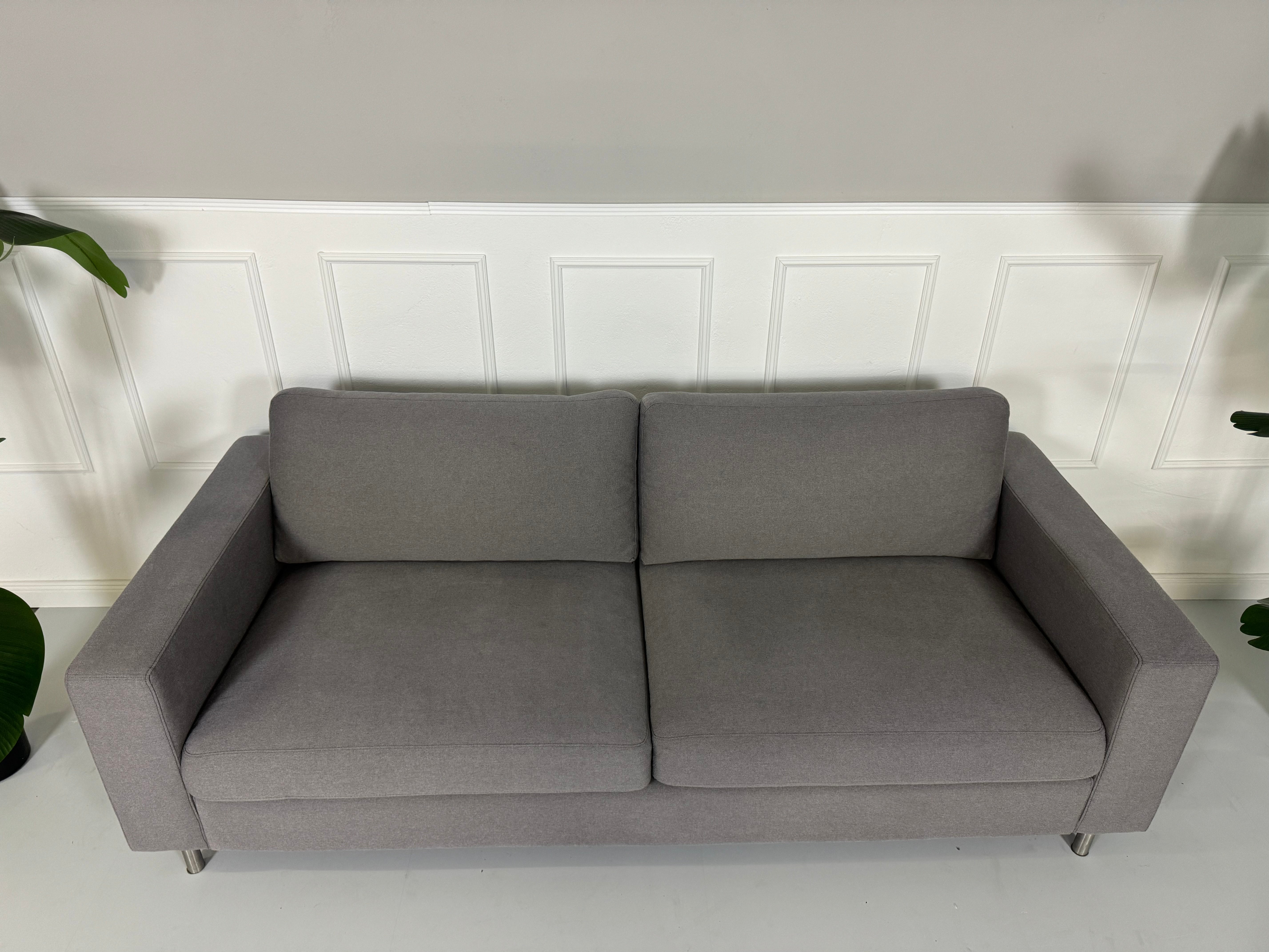 Gebrauchtes graues Bolia Scandinavia Stoff Sofa vor einer Wand, stilvoll und modern eingerichtet.