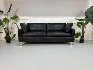 Ein gebrauchtes Sofa der Marke Brühl Modell Alba in der Farbe Schwarz Designer Leder Sofa vor einer Wand, stilvoll und modern eingerichtet.
