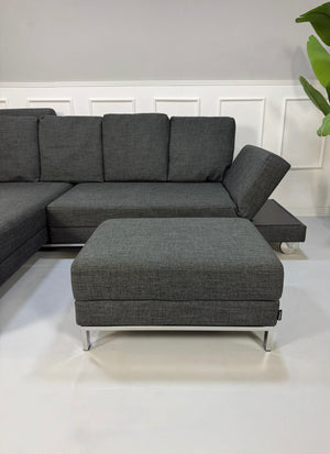 Gebrauchtes graues Brühl Fourtwo Designer Stoff Sofa mit Hocker vor einer Wand, stilvoll und modern eingerichtet.