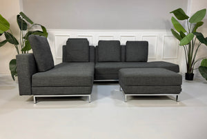 Gebrauchtes graues Brühl Fourtwo Designer Stoff Sofa mit Hocker vor einer Wand, stilvoll und modern eingerichtet.