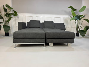 Gebrauchtes graues Brühl Fourtwo Designer Stoff Sofa mit Hocker vor einer Wand, stilvoll und modern eingerichtet.
