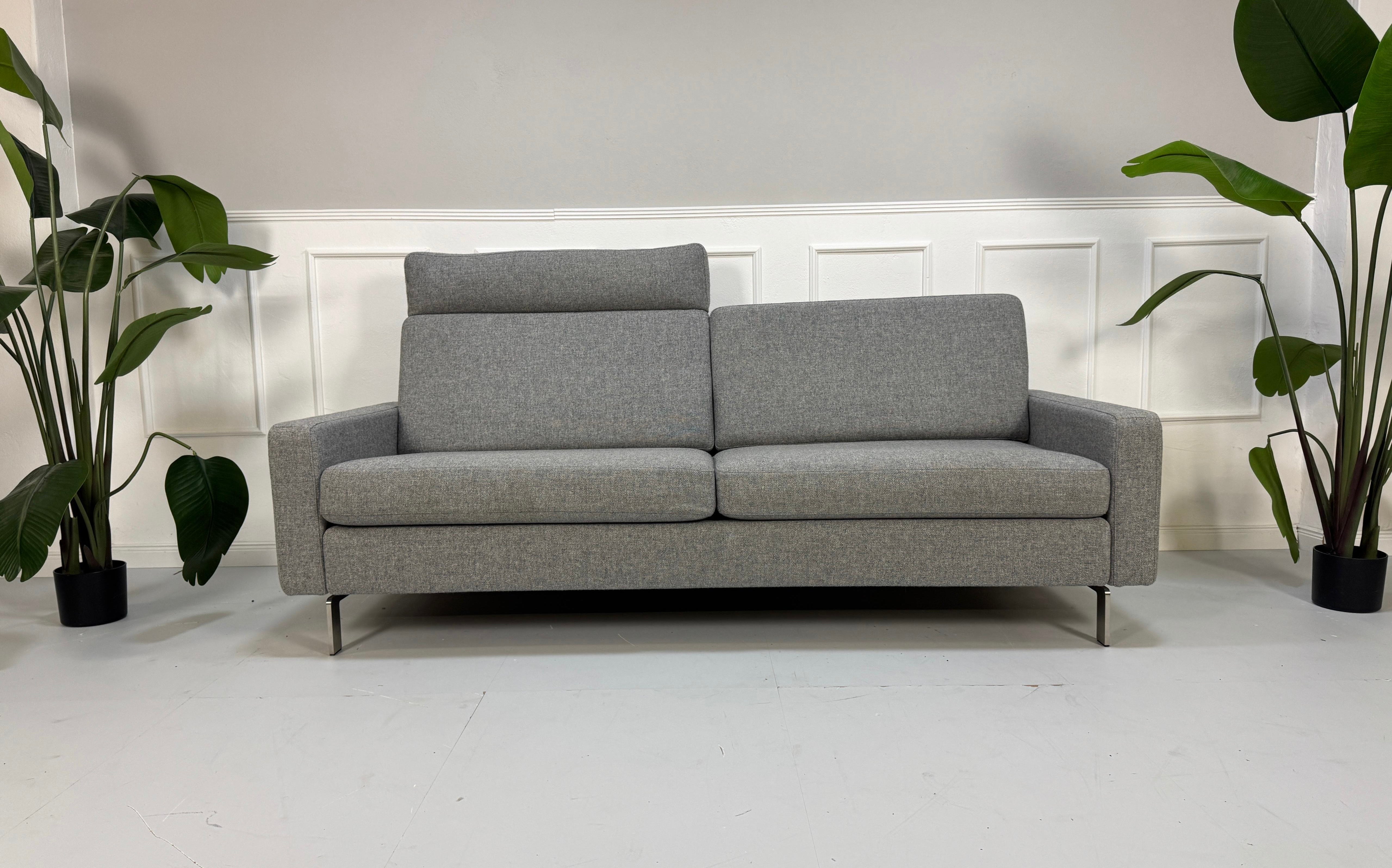 Gebrauchtes graues COR Conseta mit Kopfstütze Designer Stoff Sofa vor einer Wand, stilvoll und modern eingerichtet.