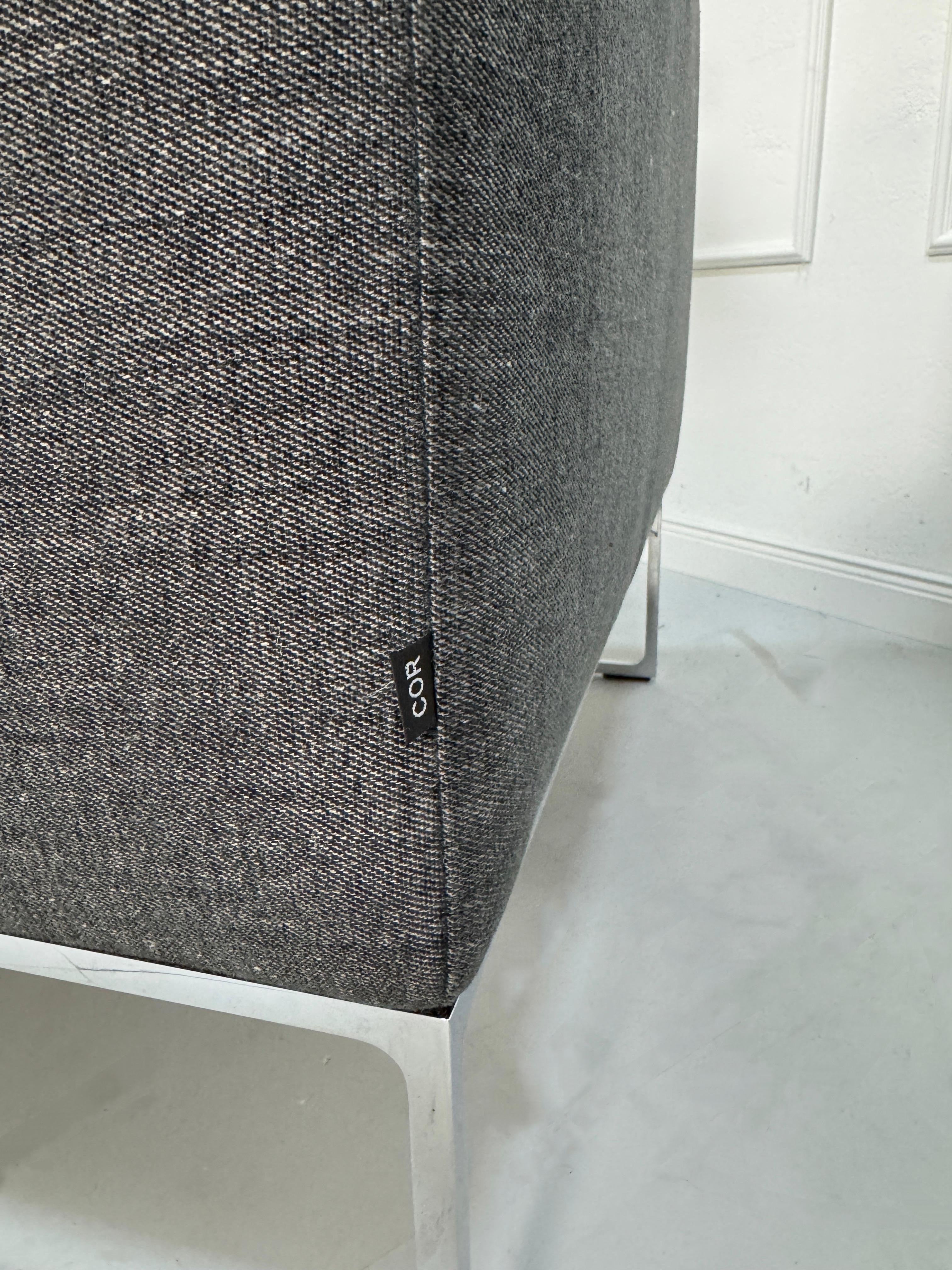 Gebrauchtes graues COR Mell Designer Stoff Sofa vor einer Wand, stilvoll und modern eingerichtet.