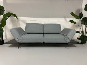Gebrauchtes grau blaues Rolf Benz Aura Designer Stoff Sofa vor einer Wand, stilvoll und modern eingerichtet.
