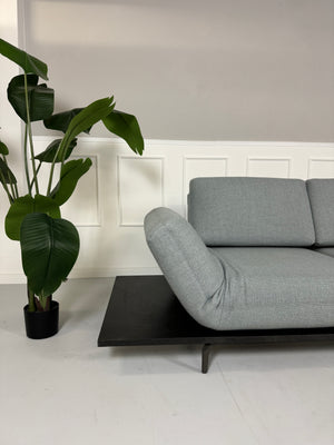 Gebrauchtes grau blaues Rolf Benz Aura Designer Stoff Sofa vor einer Wand, stilvoll und modern eingerichtet.