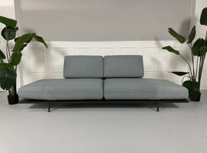 Gebrauchtes grau blaues Rolf Benz Aura Designer Stoff Sofa vor einer Wand, stilvoll und modern eingerichtet.
