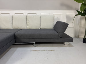 Gebrauchtes graues Brühl Fourtwo Designer Stoff Ecksofa vor einer Wand, stilvoll und modern eingerichtet.