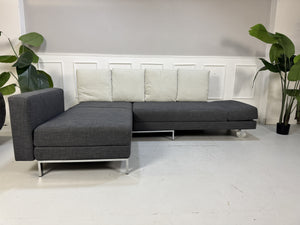 Gebrauchtes graues Brühl Fourtwo Designer Stoff Ecksofa vor einer Wand, stilvoll und modern eingerichtet.