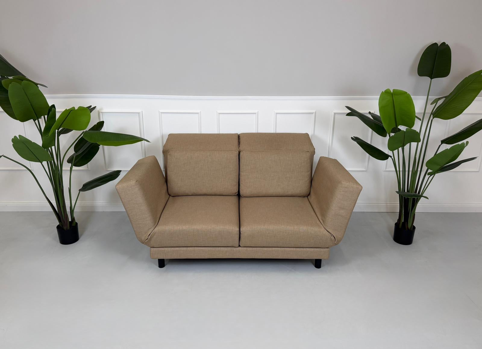 Gebrauchtes braunes Stoffsofa Brühl Modell Moule Small vor einer modernen Wand.