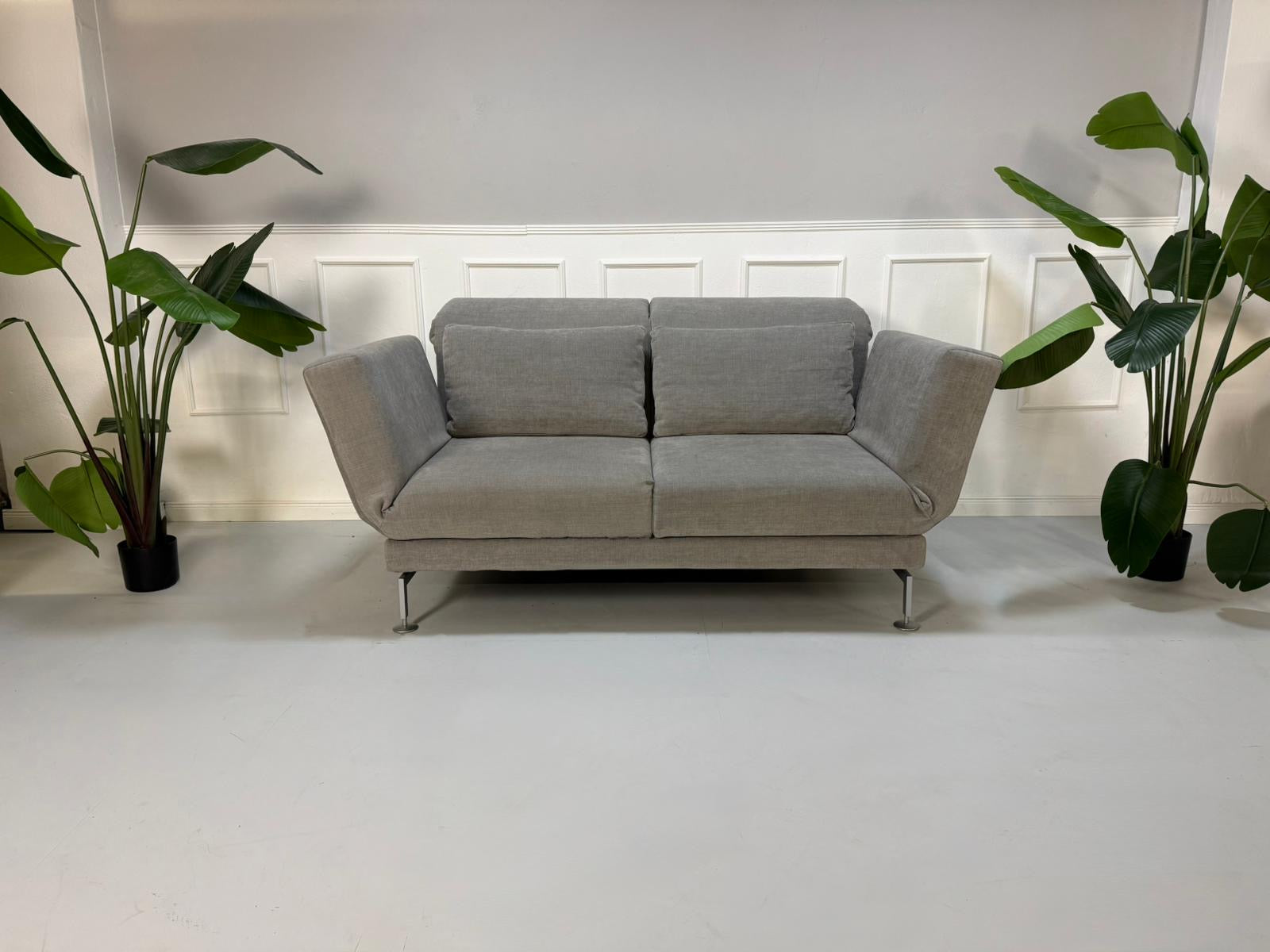 Gebrauchtes graues Stoffsofa Brühl Modell Moule vor einer modernen Wand.