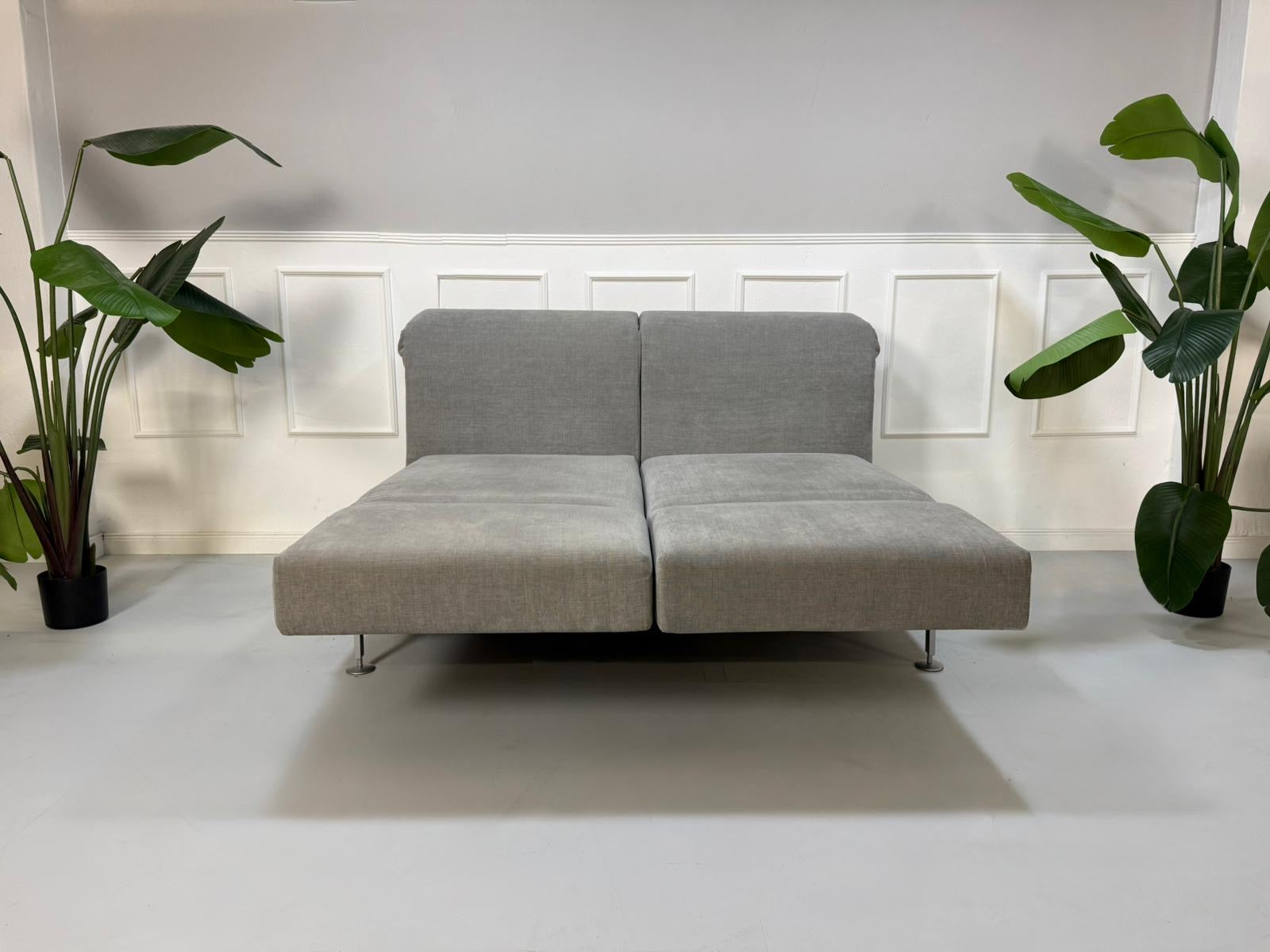 Gebrauchtes graues Stoffsofa Brühl Modell Moule vor einer modernen Wand.