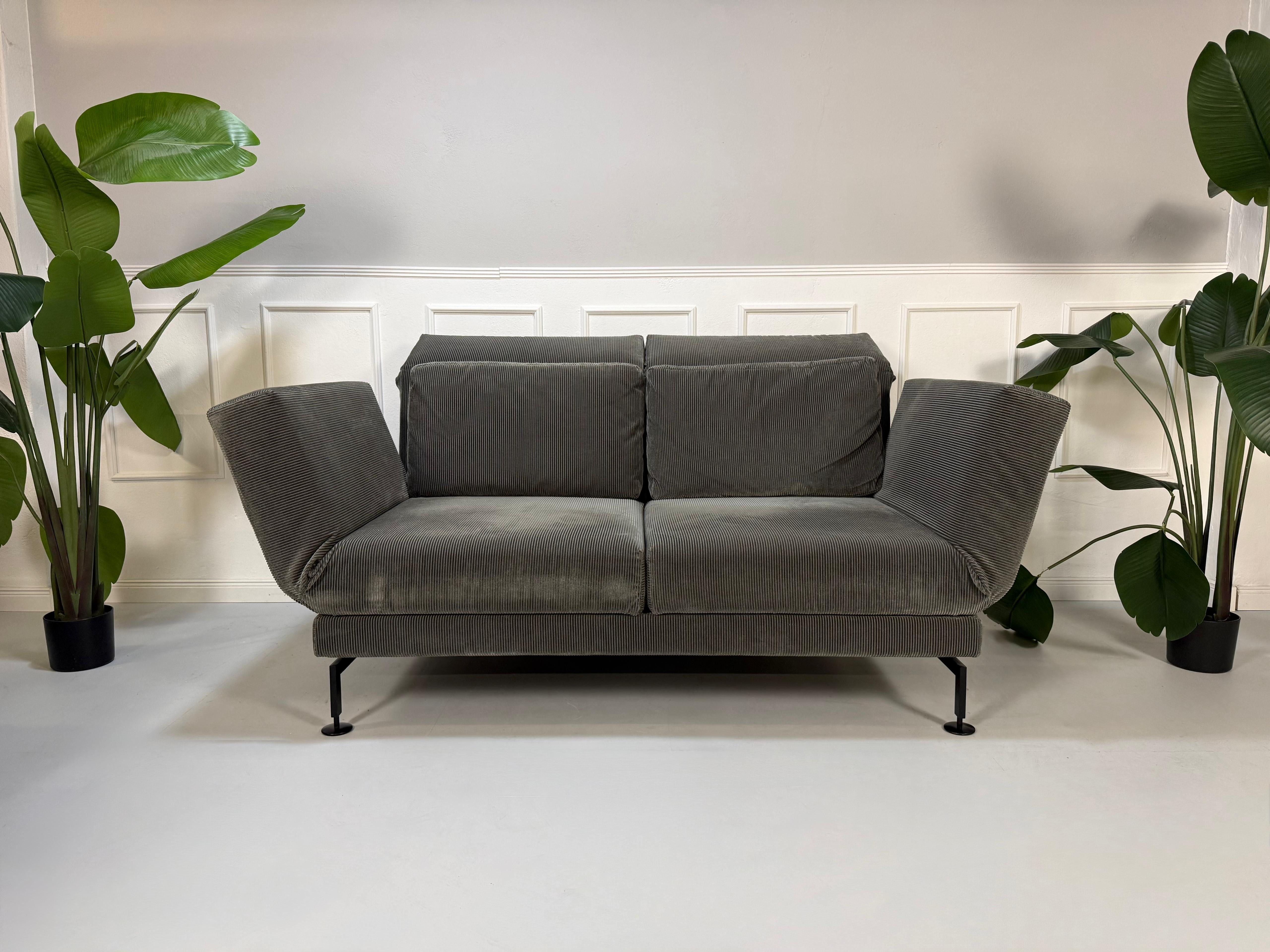 Gebrauchtes graues Kord Stoff Sofa Brühl Modell Moule Medium vor einer modernen Wand.