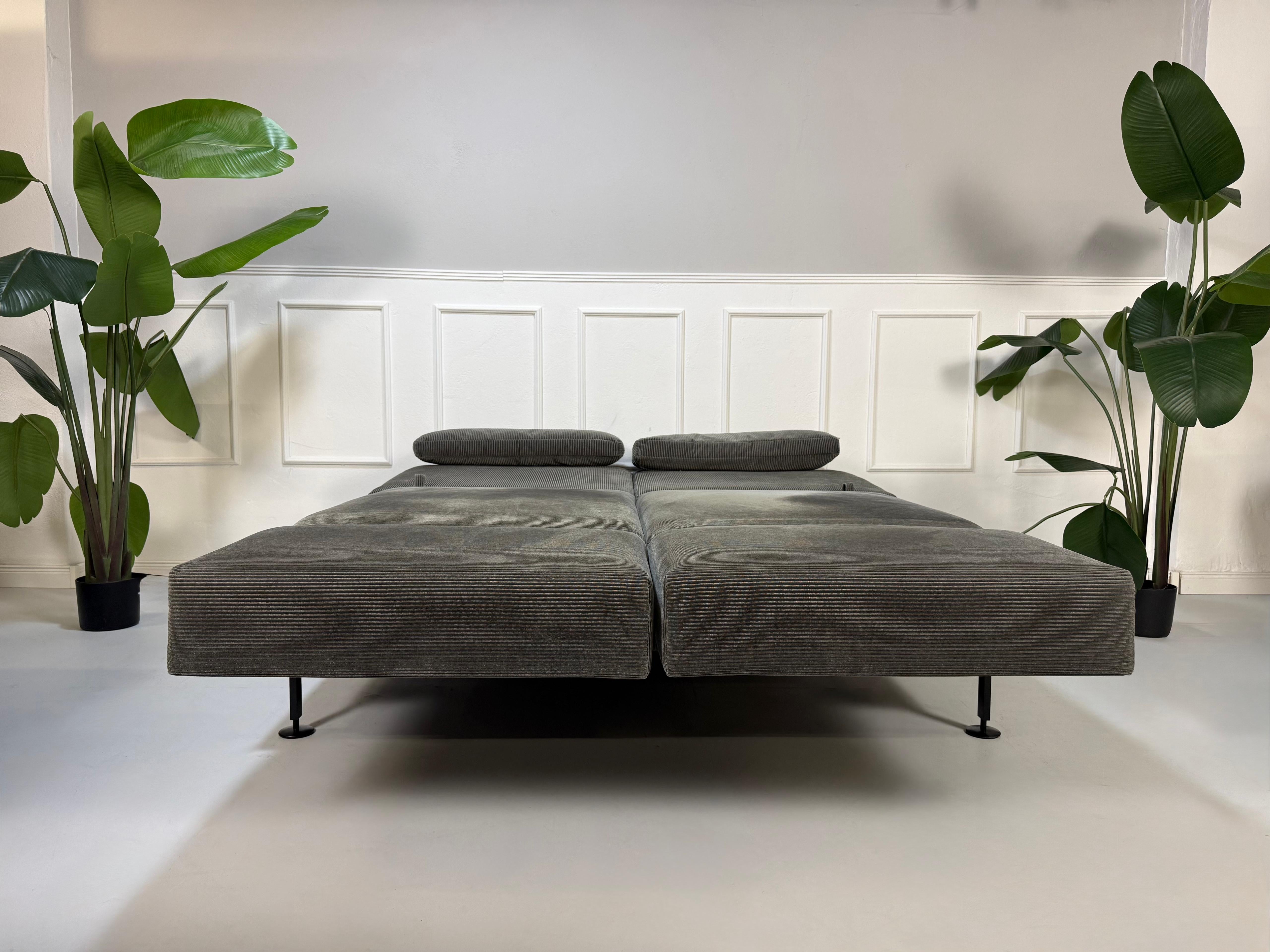 Gebrauchtes graues Kord Stoff Sofa Brühl Modell Moule Medium vor einer modernen Wand.
