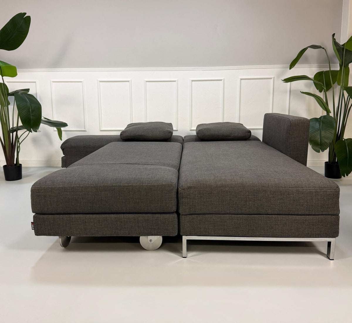 Gebrauchtes graues Brühl Fourtwo Designer Stoff Ecksofa vor einer Wand, stilvoll und modern eingerichtet.