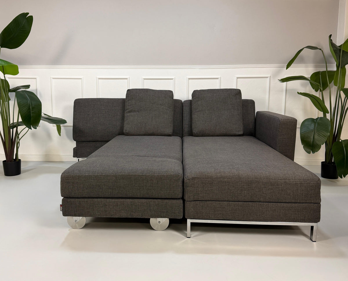 Gebrauchtes graues Brühl Fourtwo Designer Stoff Ecksofa vor einer Wand, stilvoll und modern eingerichtet.