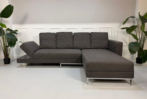 Gebrauchtes graues Brühl Fourtwo Designer Stoff Ecksofa vor einer Wand, stilvoll und modern eingerichtet.