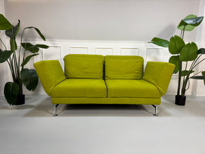 Gebrauchtes grünes Stoff Sofa Brühl Modell Moule vor einer modernen Wand.