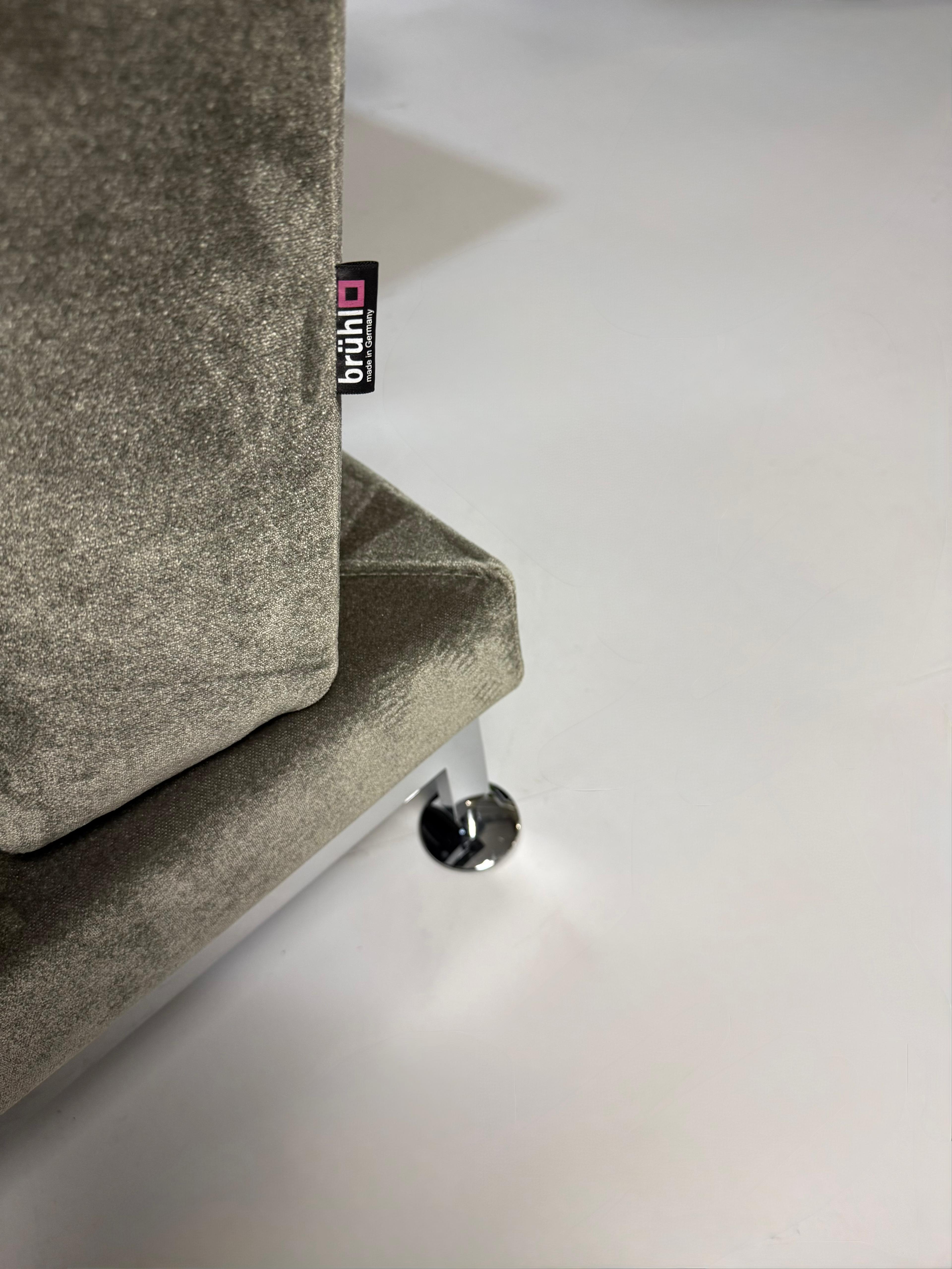 Gebrauchtes grau grünes Brühl Moule Designer Stoff Sofa vor einer Wand, stilvoll und modern eingerichtet.