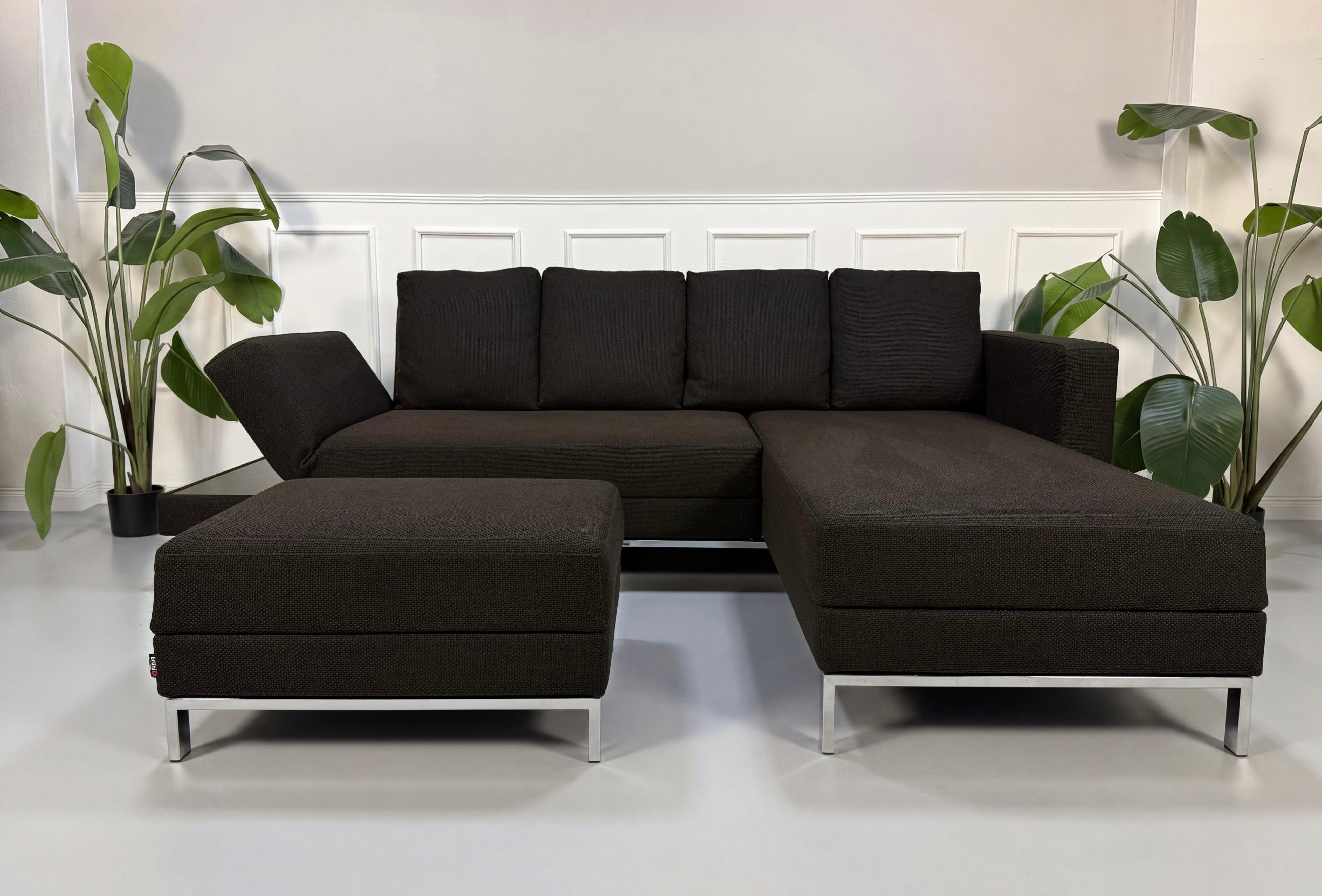 Gebrauchtes braunes Brühl Fourtwo Designer Stoff Sofa mit Hocker vor einer Wand, stilvoll und modern eingerichtet.
