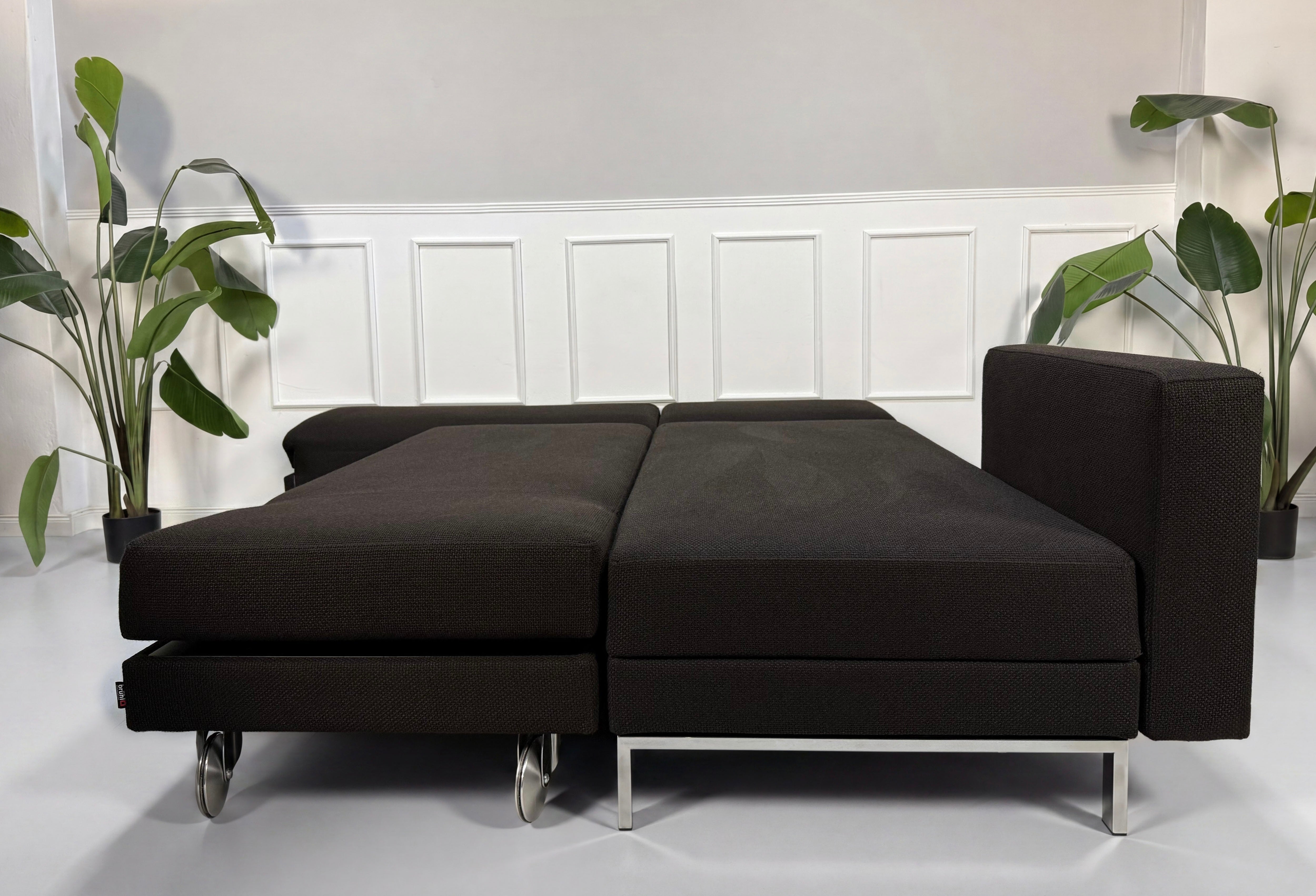 Gebrauchtes braunes Brühl Fourtwo Designer Stoff Sofa mit Hocker vor einer Wand, stilvoll und modern eingerichtet.