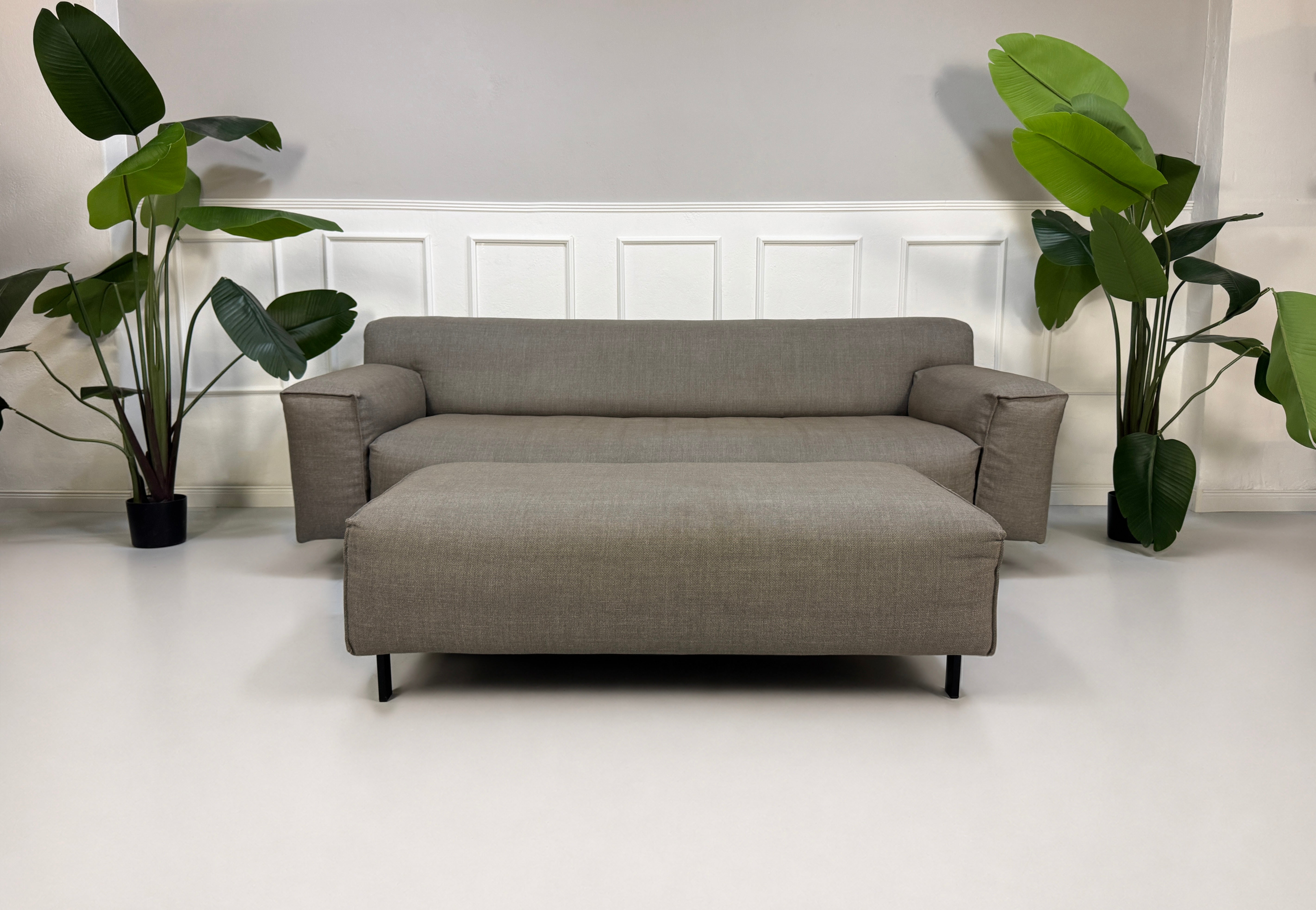 Gebrauchtes graues Rolf Benz Grata Designer Stoff Sofa mit Hocker vor einer Wand, stilvoll und modern eingerichtet.