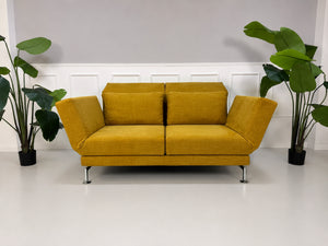 Gebrauchtes gelbes Brühl Moule Designer Stoff Sofa vor einer Wand, stilvoll und modern eingerichtet.
