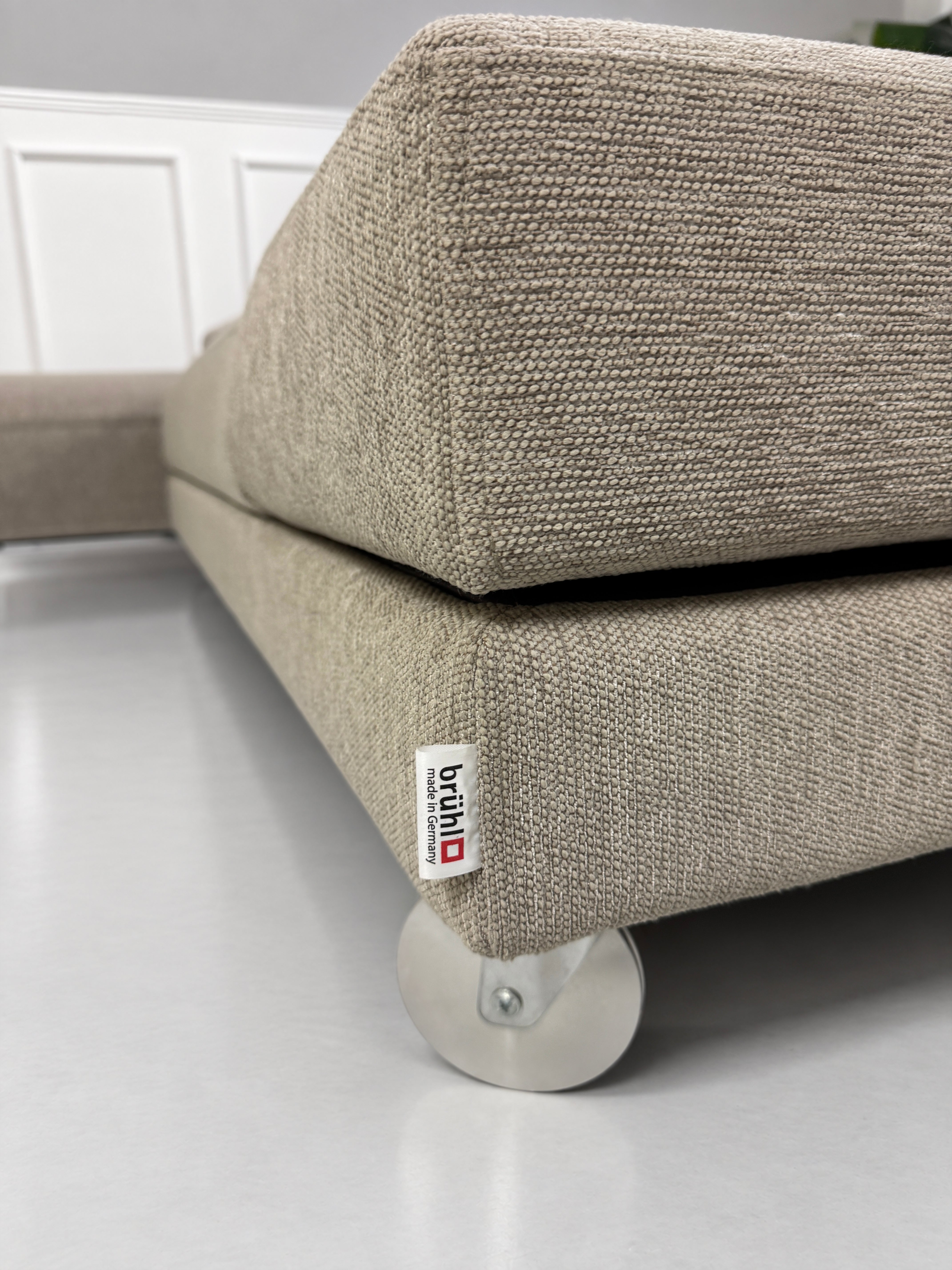 Gebrauchtes grau-beiges Brühl Fourtwo Designer Stoff Ecksofa vor einer Wand, stilvoll und modern eingerichtet.