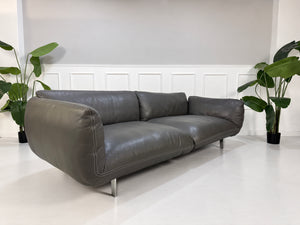 Gebrauchtes graues COR Jalis Designer Leder Sofa mit Hocker vor einer Wand, stilvoll und modern eingerichtet.