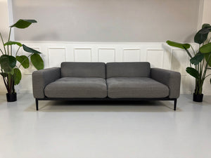 Gebrauchtes graues Rolf Benz Tira Designer Stoff Sofa vor einer Wand, stilvoll und modern eingerichtet.