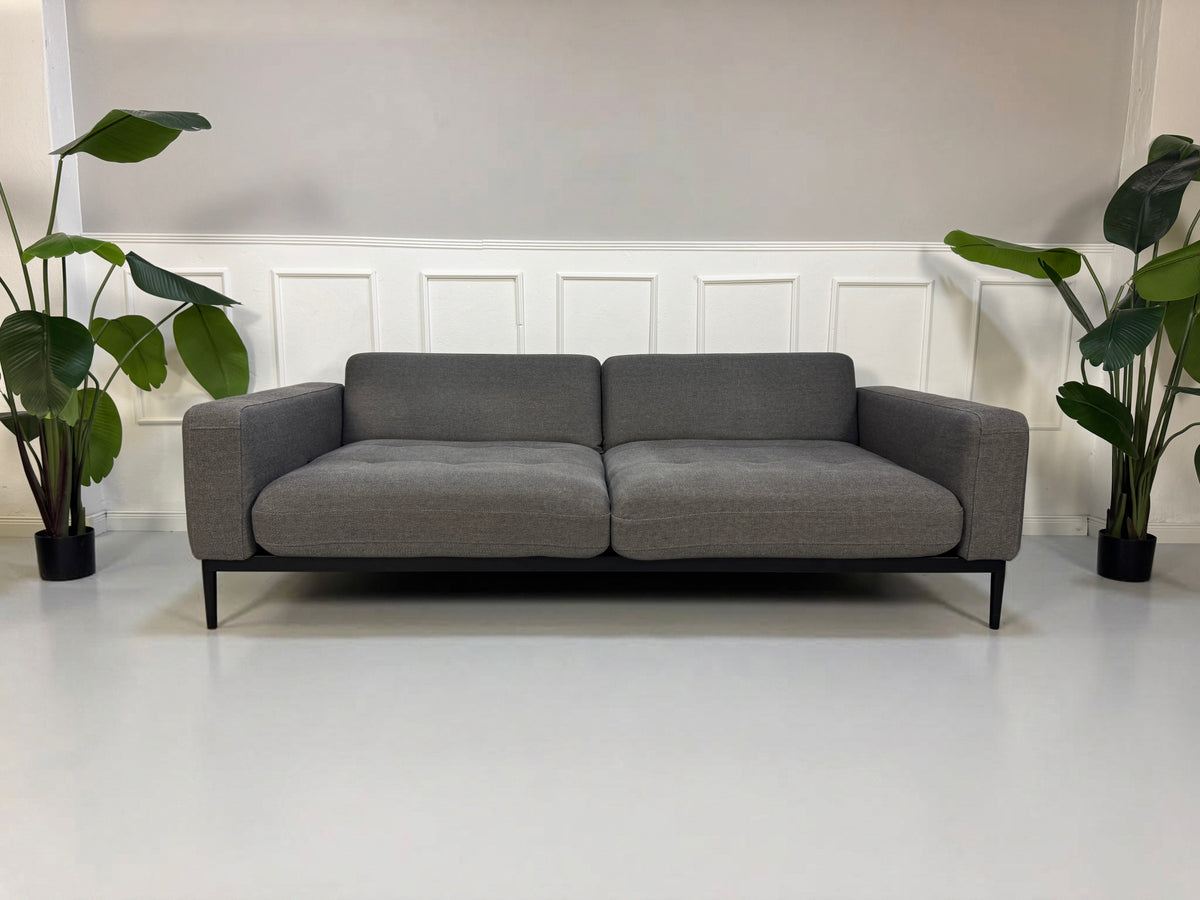Gebrauchtes graues Rolf Benz Tira Designer Stoff Sofa vor einer Wand, stilvoll und modern eingerichtet.