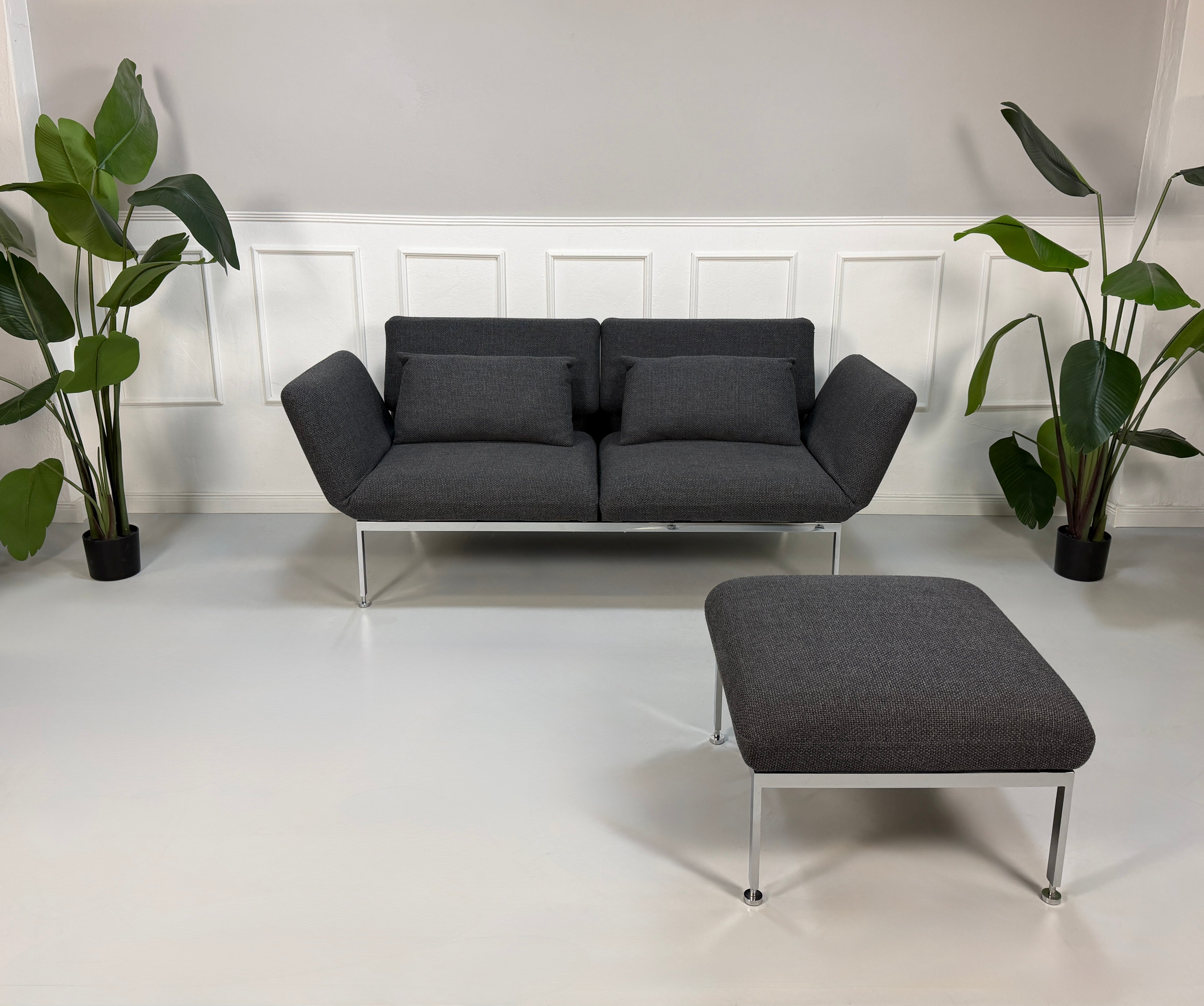 Gebrauchtes graues Brühl Roro Designer Stoff Sofa mit Hocker vor einer Wand, stilvoll und modern eingerichtet.
