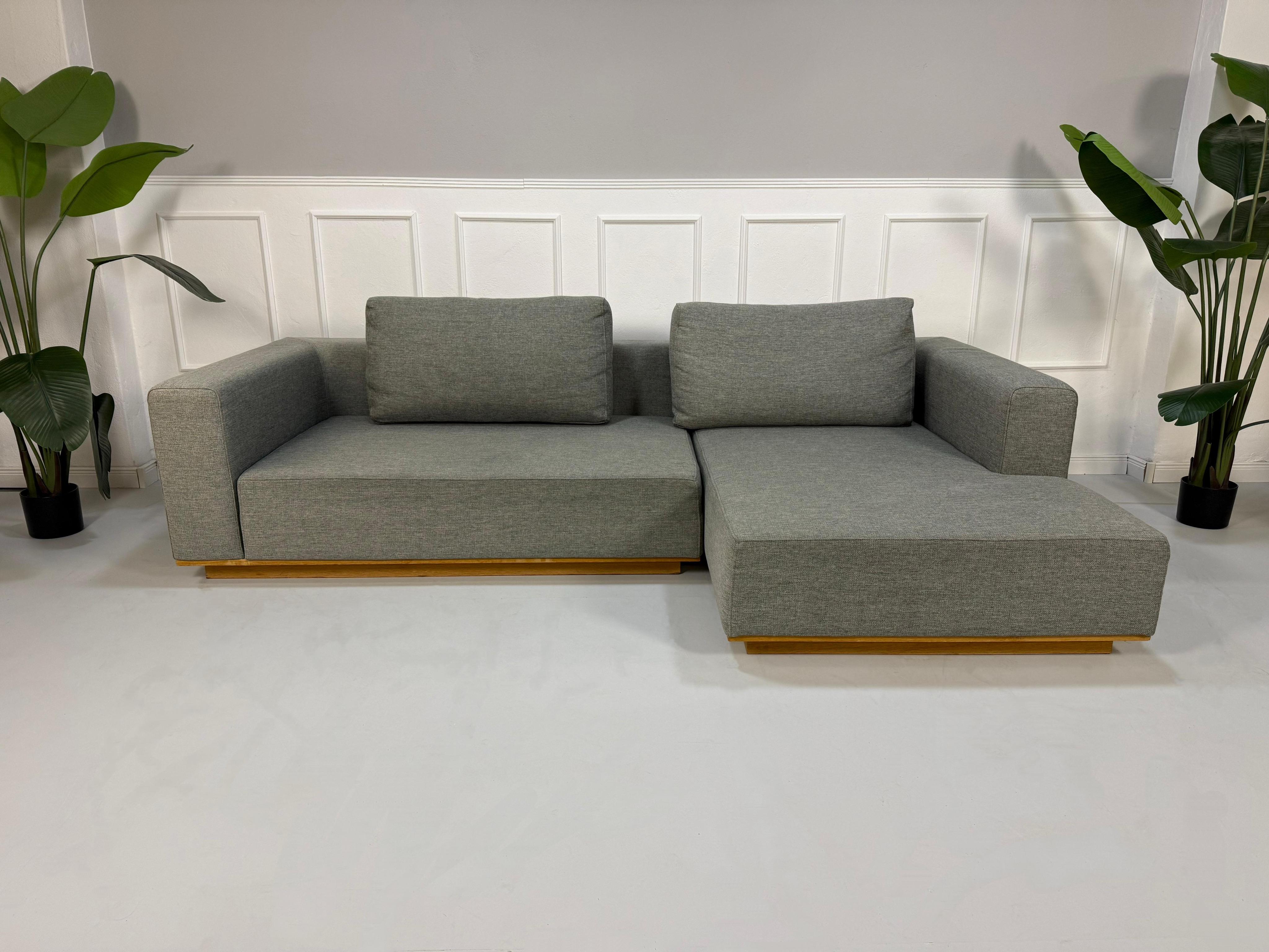 Gebrauchtes graues Bolia Element Designer Stoff Sofa mit einem Holzsockel vor einer Wand, stilvoll und modern eingerichtet.