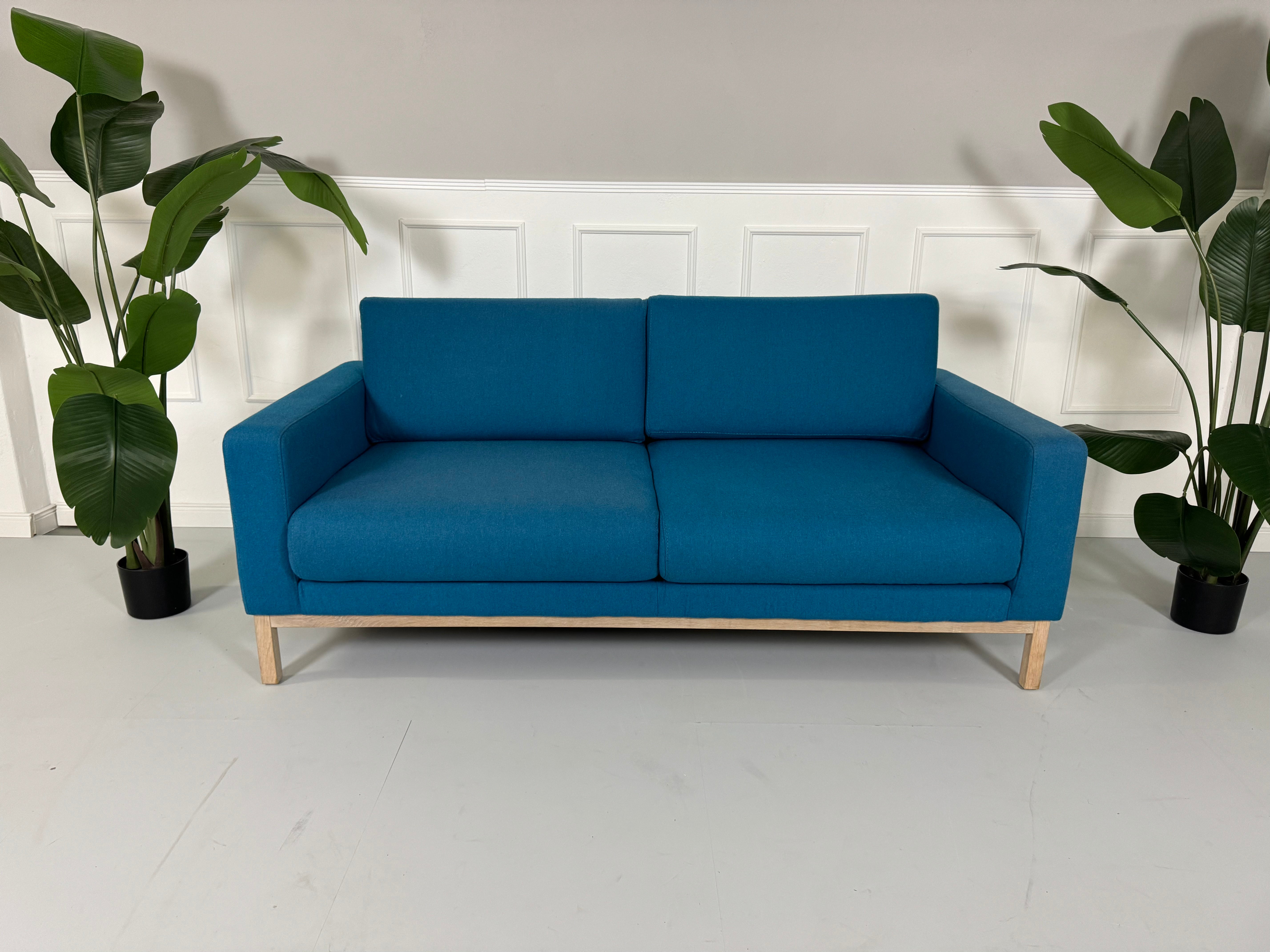 Gebrauchtes blaues petrol Bolia North Designer Stoff Sofa vor einer Wand, stilvoll und modern eingerichtet.