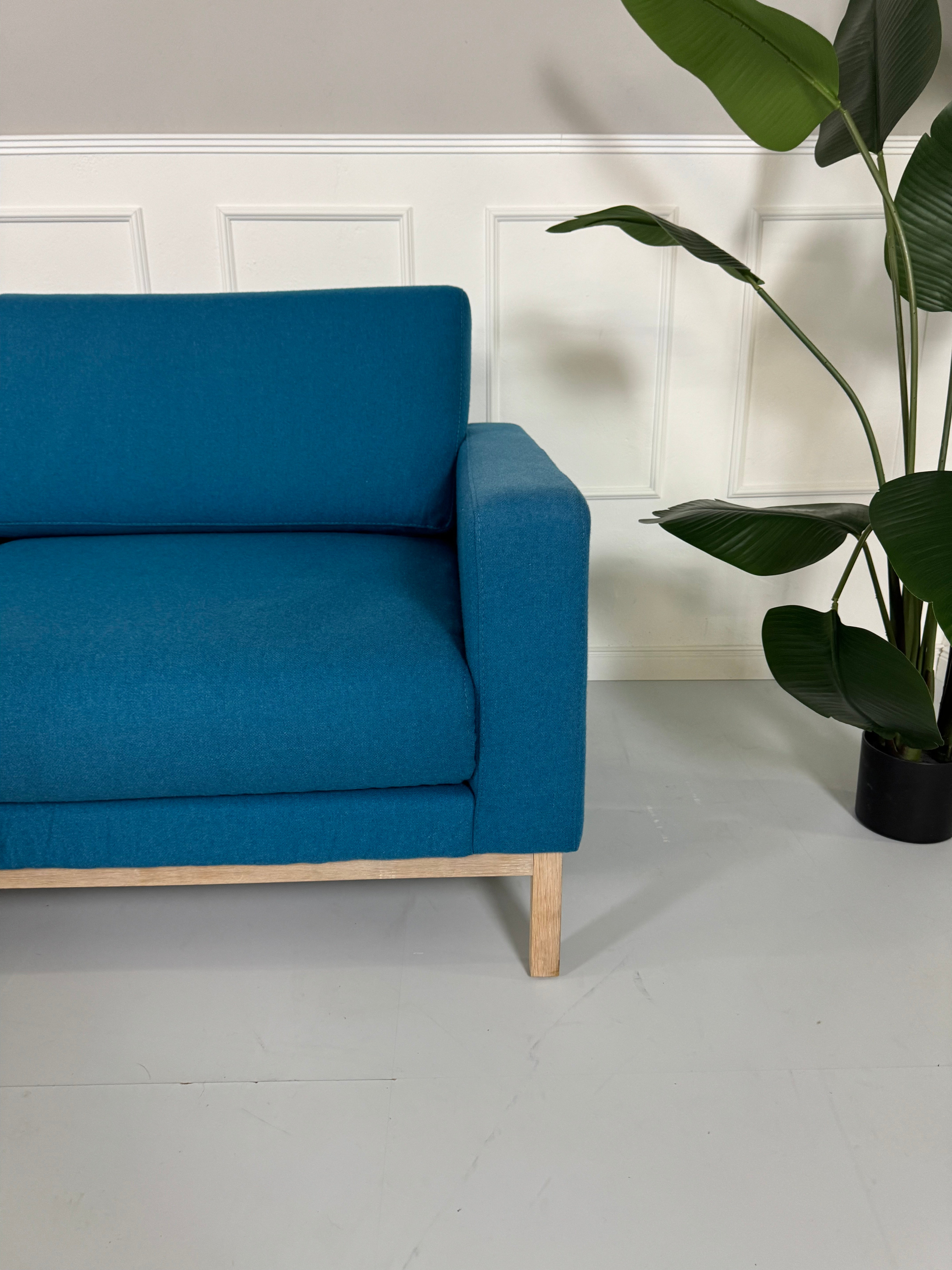 Gebrauchtes blaues petrol Bolia North Designer Stoff Sofa vor einer Wand, stilvoll und modern eingerichtet.