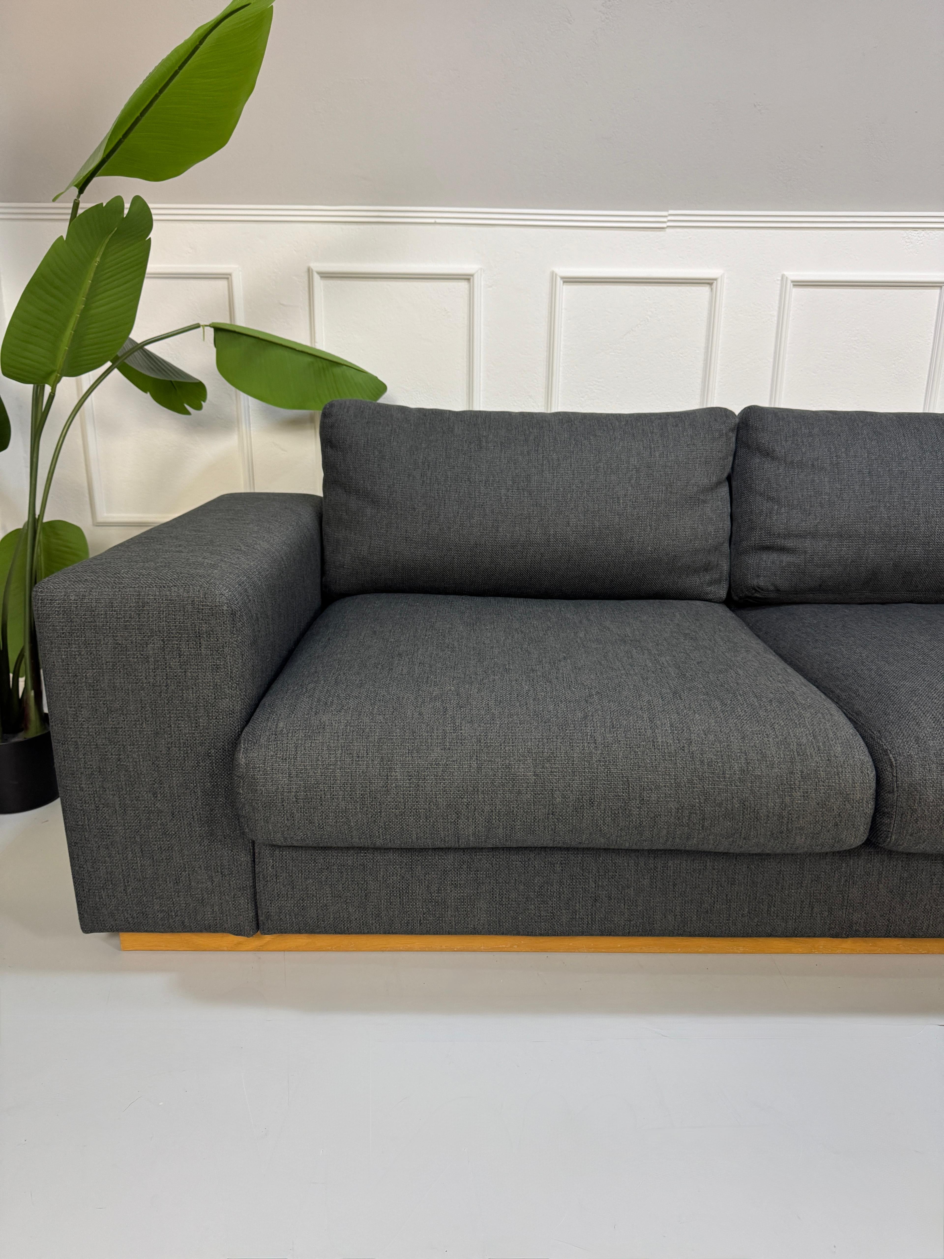 Gebrauchtes graues Bolia Sepia Designer Stoff Sofa mit Hocker vor einer Wand, stilvoll und modern eingerichtet.