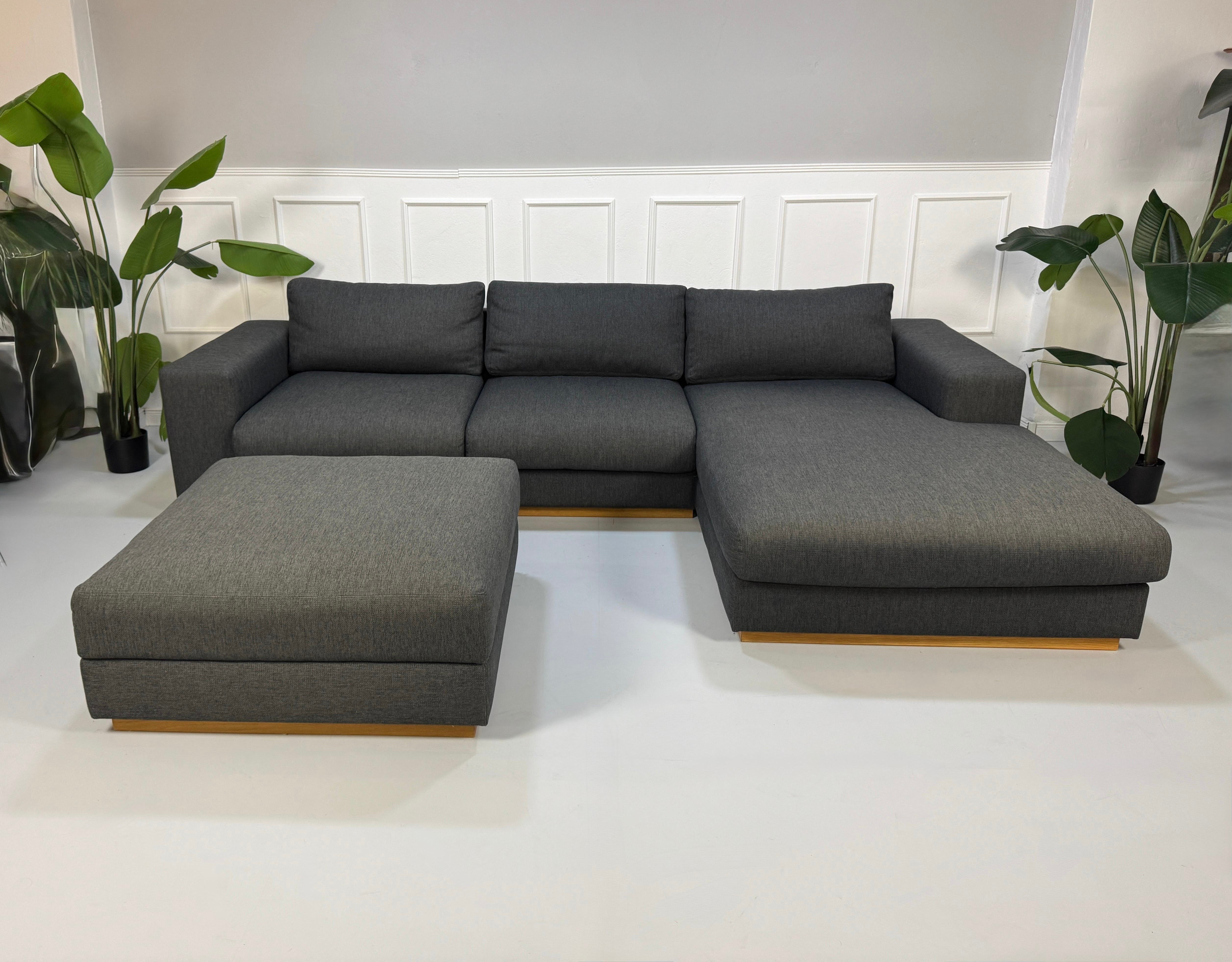 Gebrauchtes graues Bolia Sepia Designer Stoff Sofa mit Hocker vor einer Wand, stilvoll und modern eingerichtet.