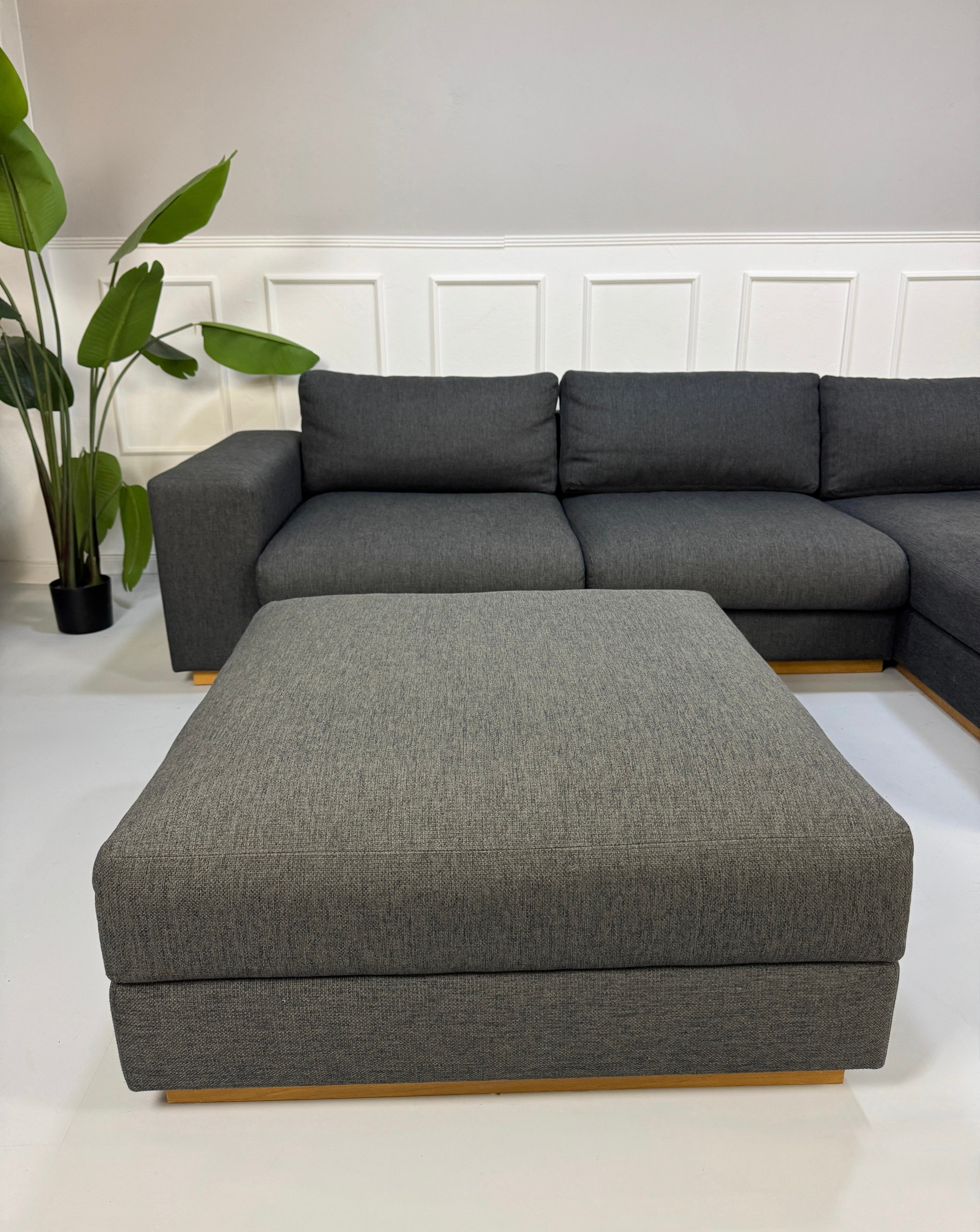 Gebrauchtes graues Bolia Sepia Designer Stoff Sofa mit Hocker vor einer Wand, stilvoll und modern eingerichtet.